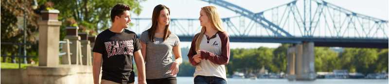 Students wearing UWL shirts walking in Riverside Park in front of the Mississippi River bridge.