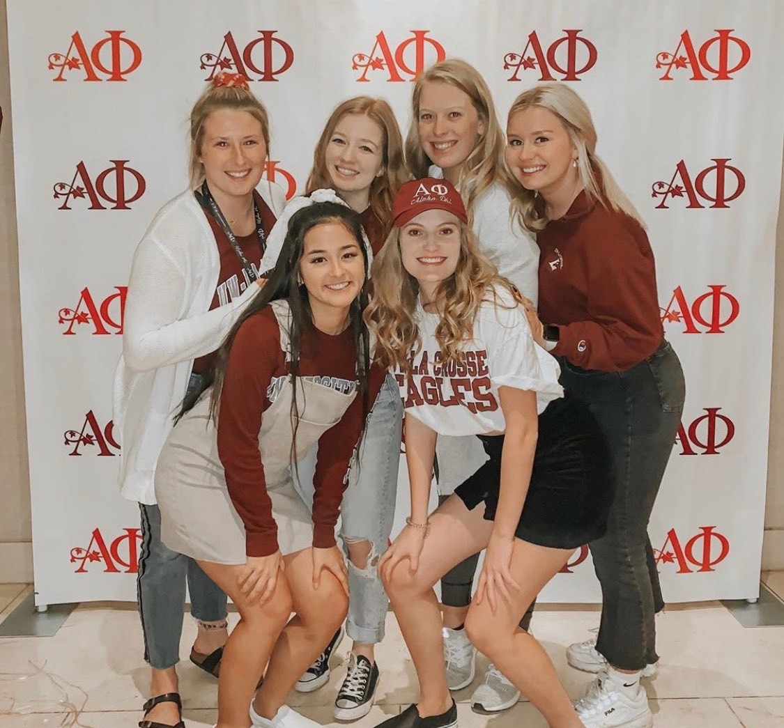 Alpha Phi members pose for a photo in front of a backdrop with their letters printed. 