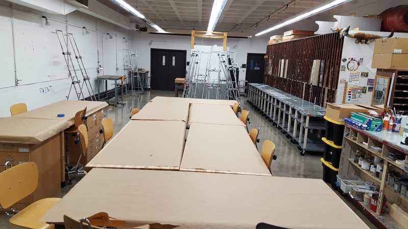 large rectangular studio with vaulted ceilings and fluorescent lights; several work tables and chairs are in the center of the space with easels on the left and in the back, and a large slat storage space on the right