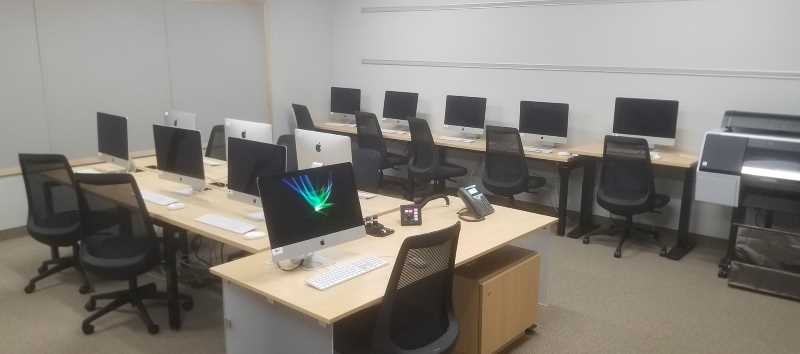 WING 27 classroom with three rows of modern light wood-top tables with several iMac desktop computers, several black mesh-back cushioned roller desk chairs, and the main desk perpendicular to the middle row of computers