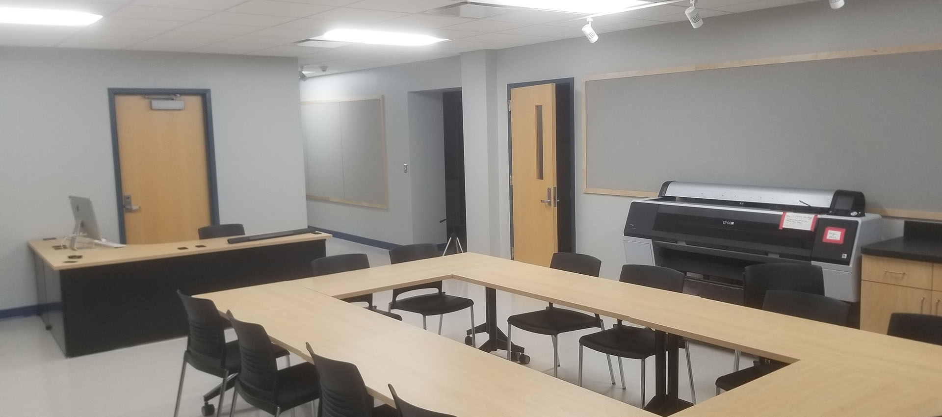 WING 27 classroom with a modern L-shaped light wood-top desk at the front, and moveable modern light wood-top tables and black hard resin chairs set up in a circle formation in the center of the room, with a large format printer in the background