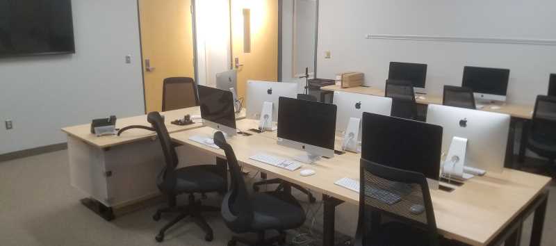 WING 28 classroom with three rows of modern light wood-top tables with several iMac desktop computers, several black mesh-back cushioned roller desk chairs, and the main desk is perpendicular to the middle row of tables