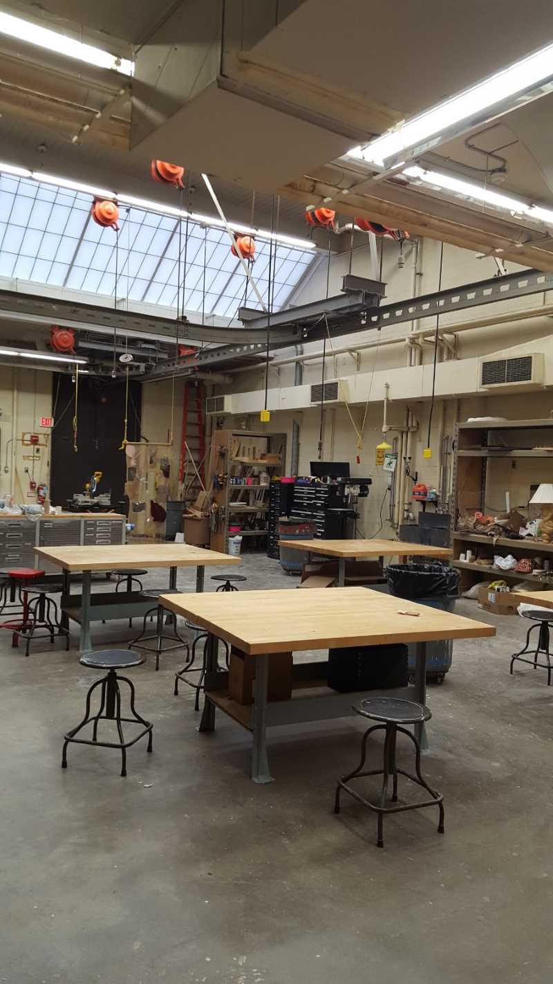 large rectangular studio with industrial vaulted ceilings and skylights; power cords hang down from a track over four large square work tables with stools; storage, tools, and table saw are in the background