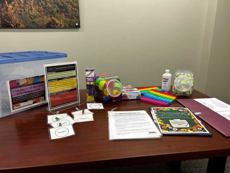 Universal design kit for programming. On the left is the plastic bin to carry the items. There are color communication badges in green, yellow, and red with a informational sign. In the middle of the image are the fidgets, accessibility checklist, and food allergy labels. On the right of the image are hand sanitizer, ear plugs, and coloring sheets. 