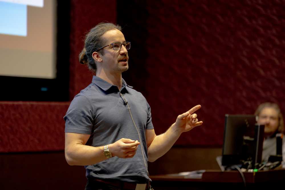 Person speaking with a microphone on stage, gesturing with their hand.