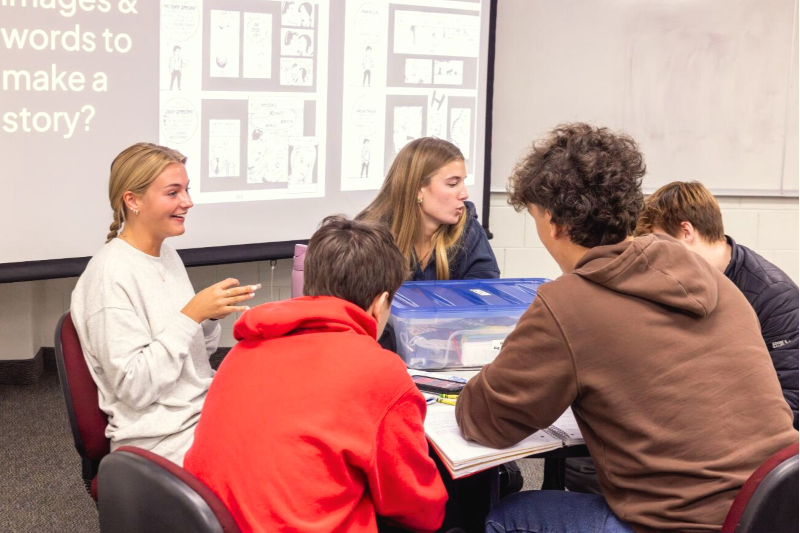 Alt text: Five HIS 110 students talking to each other at a table in a zine-based literacy instruction session. Zine creation materials are on the table.