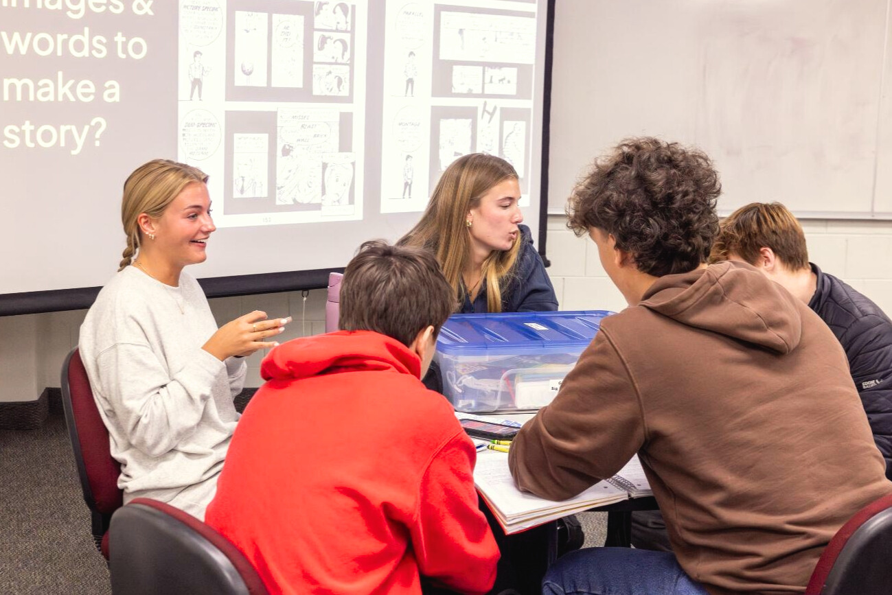 Alt text: Five HIS 110 students talking to each other at a table in a zine-based literacy instruction session. Zine creation materials are on the table.