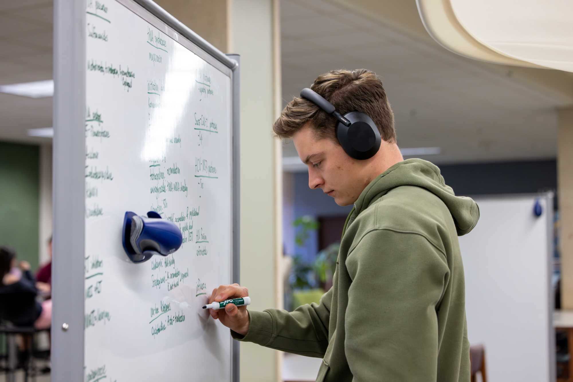 Alt text: Student in green sweatshirt writes on moveable whiteboard in Murphy Library.