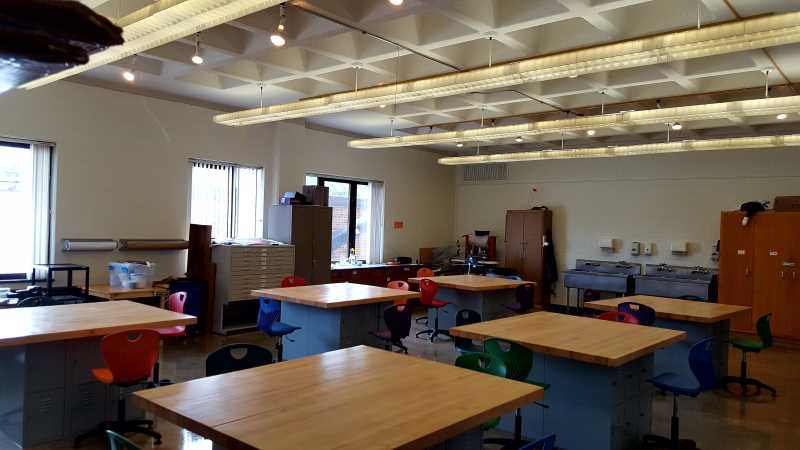 large rectangular studio with vaulted ceilings and fluorescent lights; plenty of natural light streams in from North-facing windows onto several counter-height work tables with storage lockers underneath and stools surrounding each table; there are two large sinks and storage cabinet in background