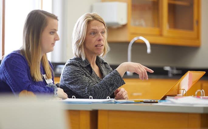 Leslie Rogers discussing with students at a table. One other student is visible in the photo.