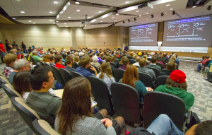 Image of Adam Reiss speaking to a large crowd in an auditorium at UW-L.