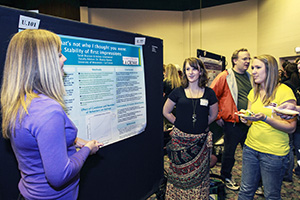 Image of a student standing by her research poster while others look on.