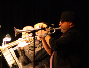 Image of a man playing a trombone. Other individuals playing musical instruments are in the background. 