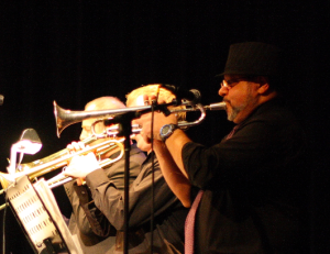 Image of a man playing a trombone. Other individuals playing musical instruments are in the background.