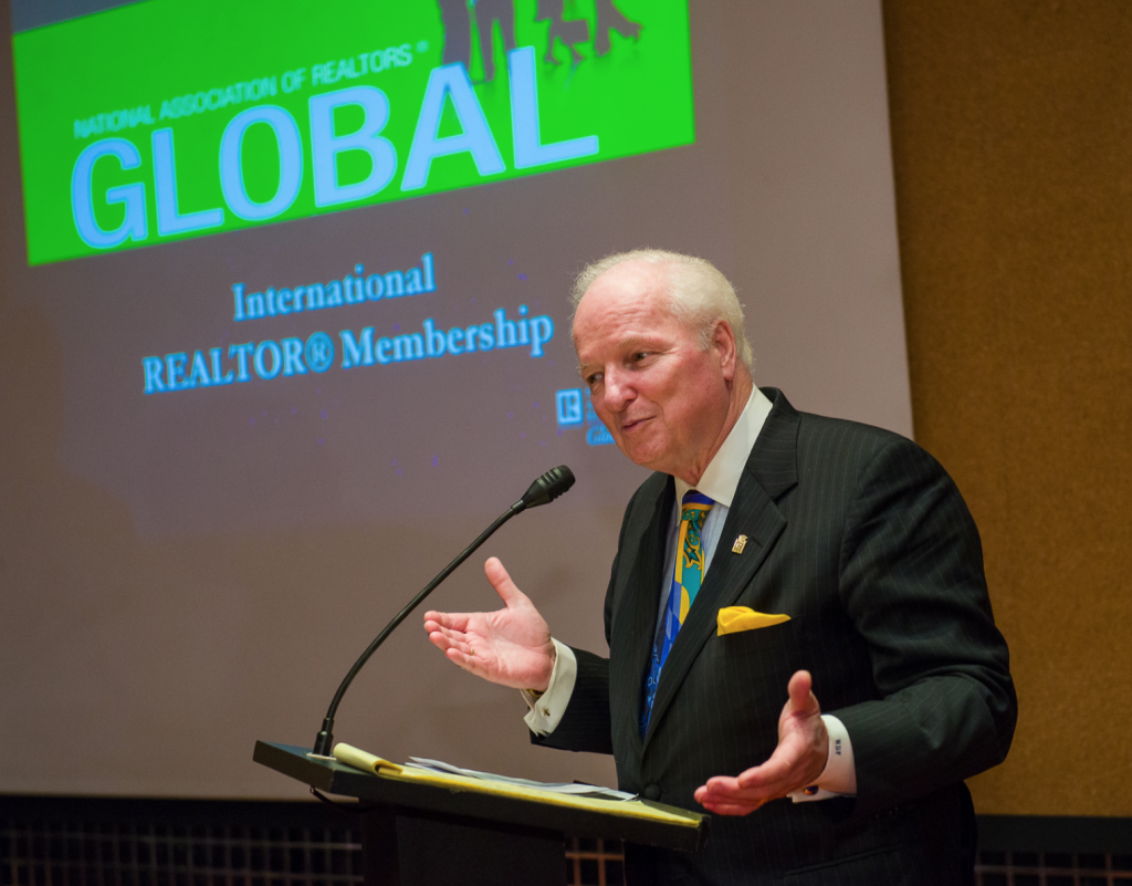 Norm Flynn speaking in front of a podium with lecture slides at a conference. 