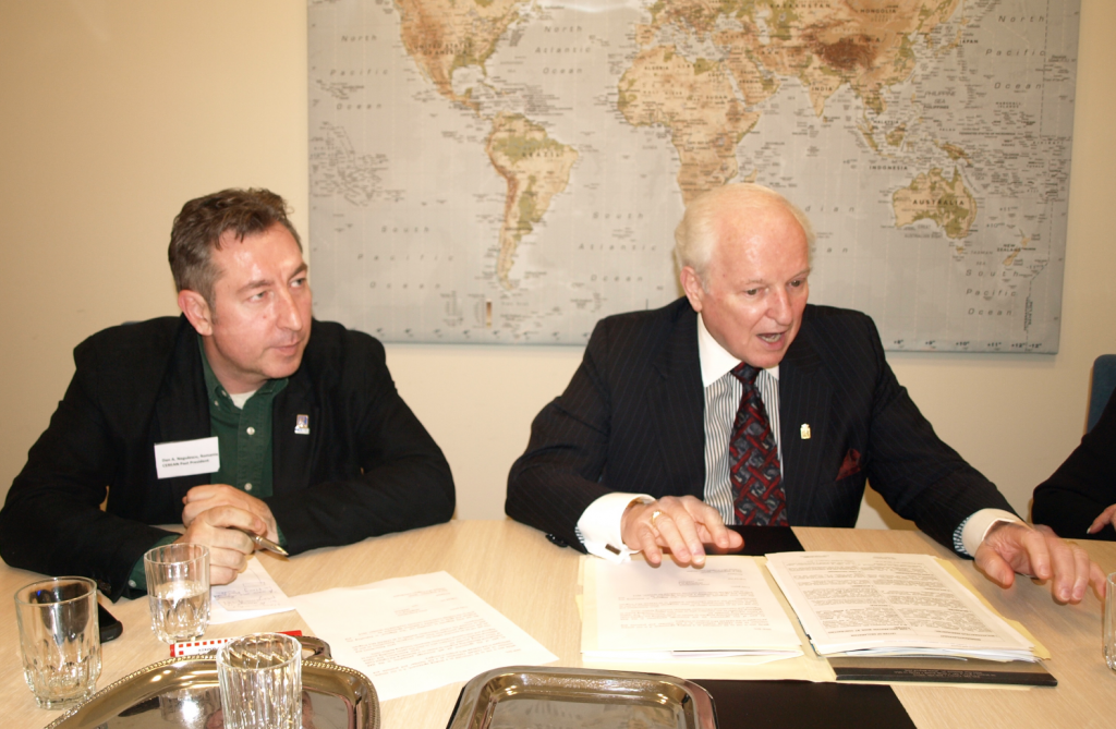 Norm Flynn sitting at a table talking with another man. World map is in the background.