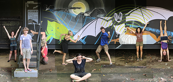 Image of people in artistic poses standing in front of a mural with painted bats, insects and a frog.