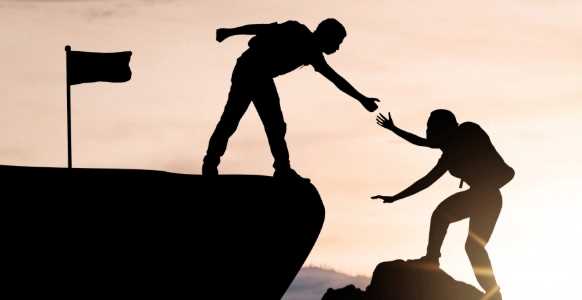 Two climbers on a mountain, with one helping the other up near the summit.