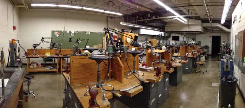 view of metals studio from front of room; vaulted ceiling with fluorescent lighting;18 solid wood-top workstations with individual vices, work lamps, and stools