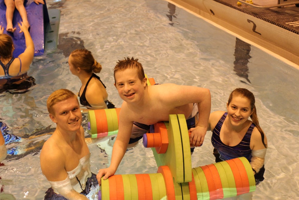 A boy with a disability rides on a floating motorcycle toy in the pool with help from Adapted Physical Education Teaching Minors.