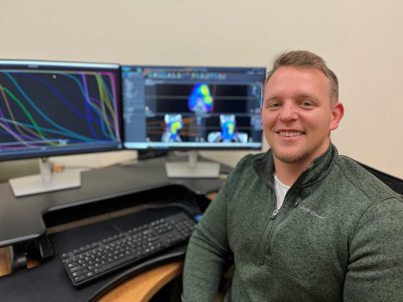 Medical Dosimetry program's alum, Jacob Wudtke, sitting at a desk at work, with two monitors displaying complex graphics, including wavy lines and heat map-like visuals.