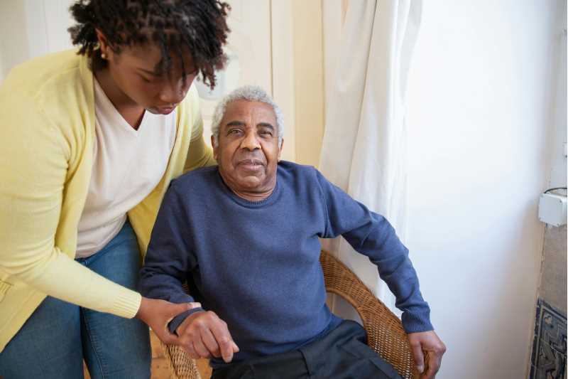 A young woman in a yellow cardigan helps an elderly man in a navy sweater to get up from a sitting position.