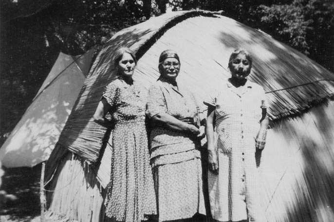 Ethel Wilson, Nettie Wapp and Nellie Wabiness standing in front of a wigwam covered with woven mats.