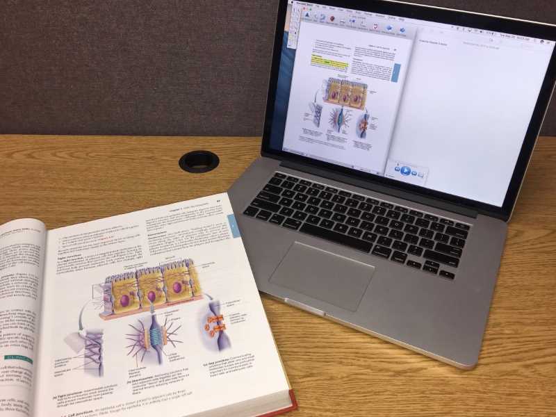 Photo of a laptop on a desk with text to speech software. A textbook is on the corner of the laptop. 