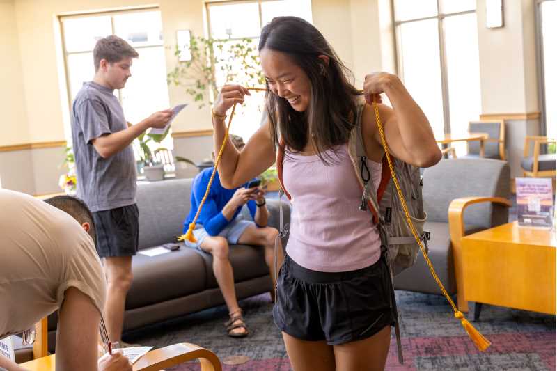 A young woman in a pink tank top and black shorts smiles while holding a yellow cord from her backpack in a university lounge, with two men engaged in conversation behind her.