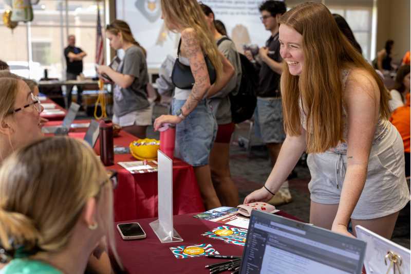 A lively university event with students engaging at tables, featuring a smiling woman in the foreground. Various promotional materials and personal items are visible on the table.