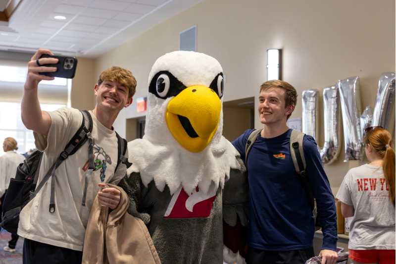 Two students take a selfie with a large eagle mascot in an indoor setting, smiling and posing together.